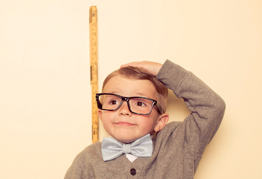 Young boy in black rimmed glasses measuring his own height with a tape measure.